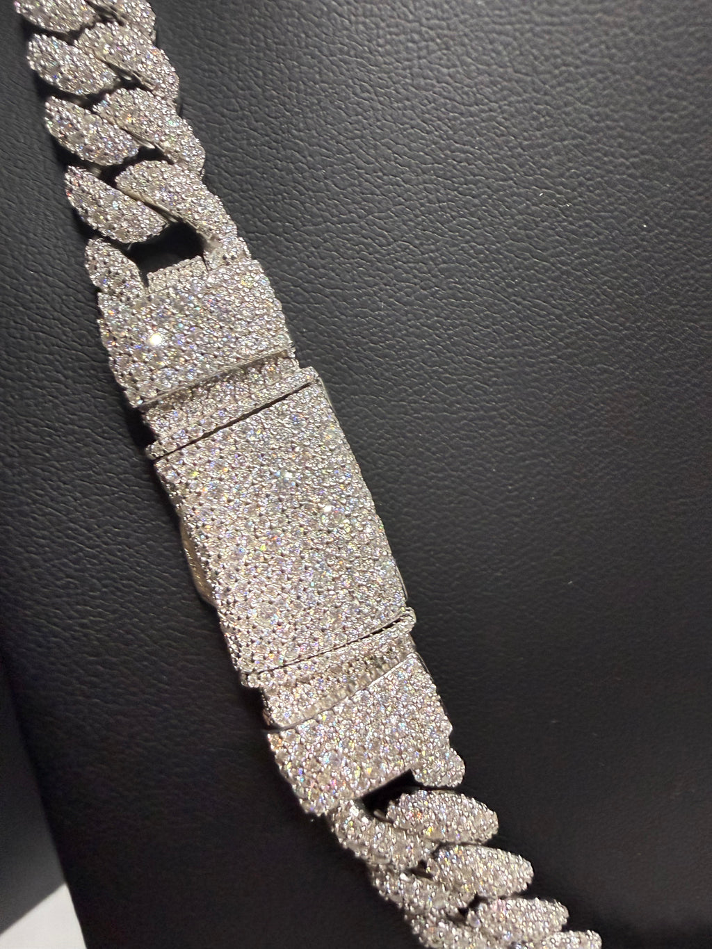 A close-up of a thick Cuban link chain bracelet covered in sparkling clear stones, displayed diagonally on a textured black surface.