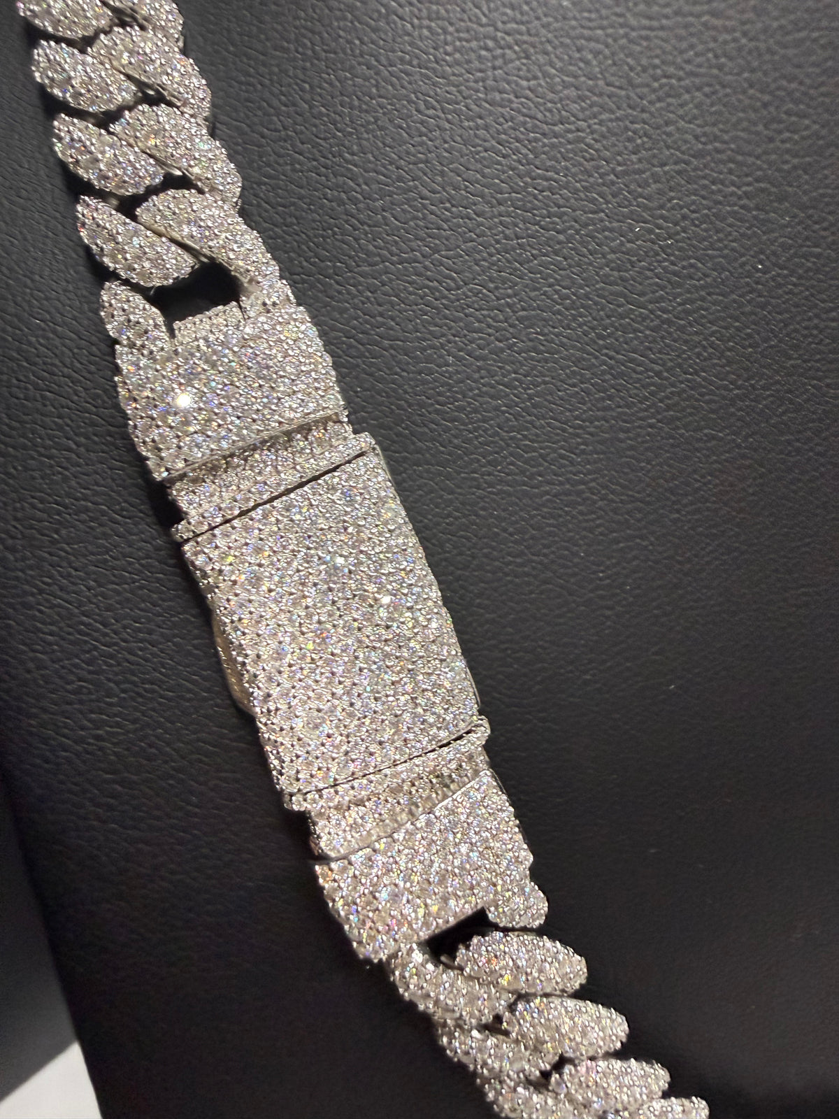 A close-up of a thick Cuban link chain bracelet covered in sparkling clear stones, displayed diagonally on a textured black surface.